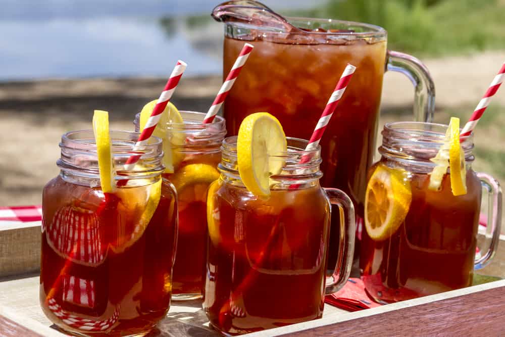 Pitcher of tea and four glasses of tea with straws and lemon wedges.