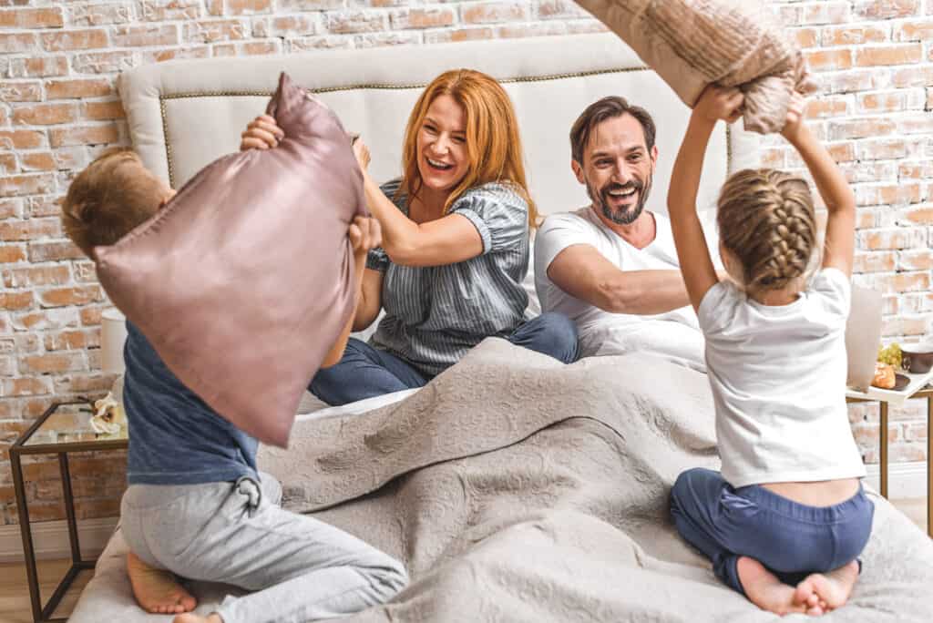 Morning battle. Photo of adult happy parents playing with their little children with pillows while staying in bed at home