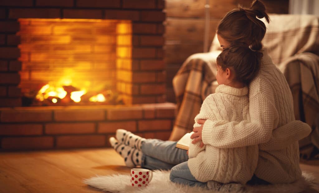 Mother and child hugging in front of a fireplace. Mom has a book on her lap and a mug is on the rug next to the child.